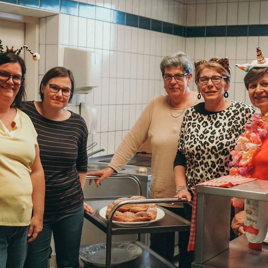 Fünf Frauen stehen in einer Küche, tragen Partyhüte und Brillen. Sie lächeln und posieren für ein Foto. Ein Tablett mit Essen und eine Ketchupflasche sind auf einem Trolley. Ein Waschbecken und ein Spender sind im Hintergrund.