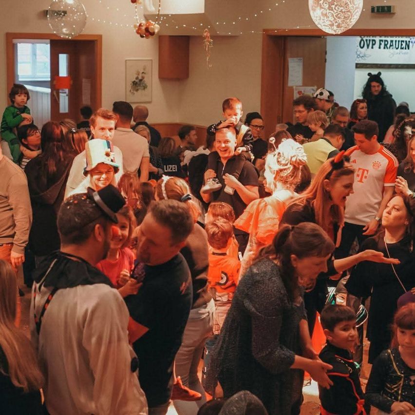 Ein überfüllter Raum mit Menschen, einige in Kostümen, drinnen mit Dekorationen, einschließlich einer Person in einem grünen Shirt, die ein Kind hält.