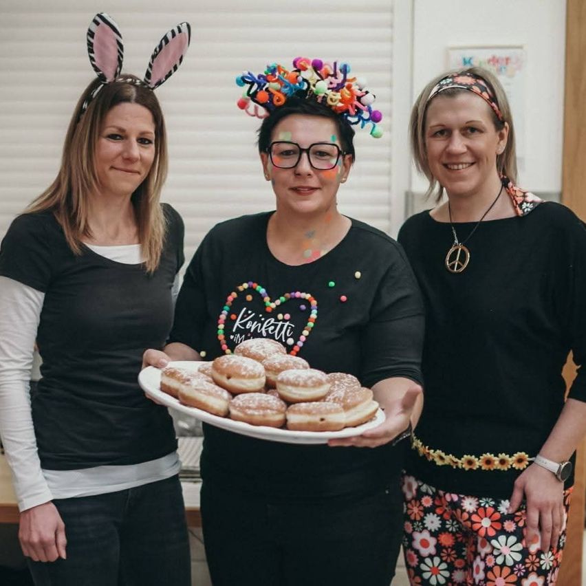 Drei Frauen in Schwarz gekleidet, eine mit Hasenohren, eine mit einer bunten Kopfband und eine mit einer Halskette, stehen nebeneinander. Die Frau in der Mitte hält einen Teller mit Donuts.