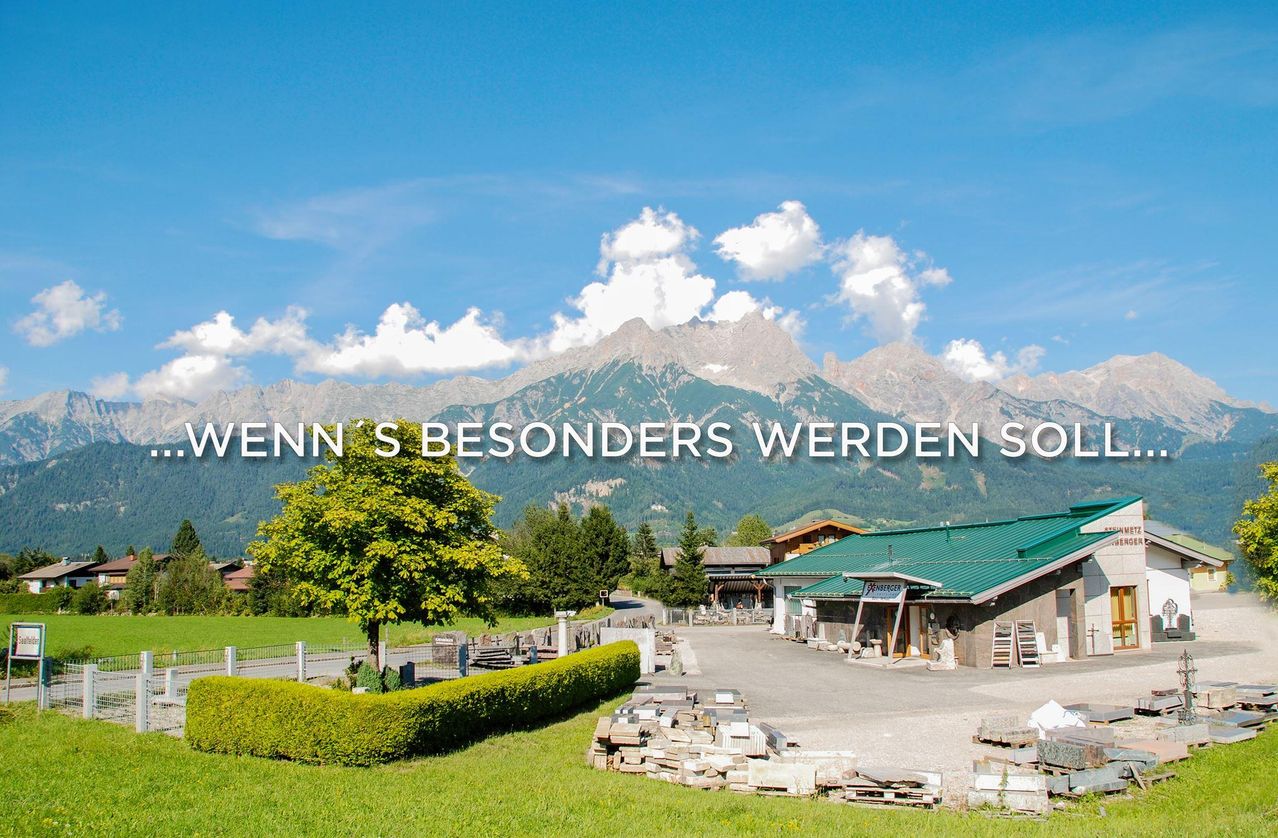 A scenic view of a mountain with a green building, a tree, and a fence. There are rocks and a hedge in the foreground. The sky is clear with clouds.