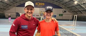 Zwei Männer posieren mit Tennisschlägern und Pokalen auf einem Tennisplatz. Einer trägt eine rote Kapuzenjacke, der andere ein rotes T-Shirt. Sie stehen auf einem Platz mit einem Netz im Hintergrund.