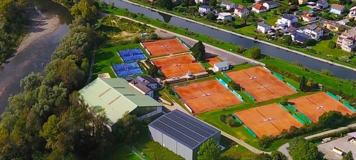 Eine Luftaufnahme einer Gemeinde mit Tennisplätzen, Häusern und einem Fluss. Das Gebiet hat grüne Felder, Bäume und einen Gehweg.