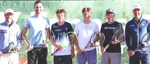 Sechs Männer stehen auf einem Tennisplatz und halten alle Tennisschläger. Sie tragen Sportkleidung und lächeln für ein Foto. Tennisbälle liegen verstreut auf dem Boden vor ihnen.