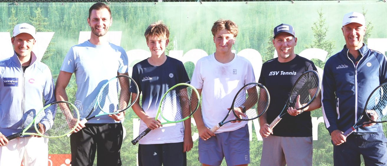 Sechs Männer stehen auf einem Tennisplatz und halten alle Tennisschläger. Sie tragen Sportkleidung und lächeln für ein Foto. Tennisbälle liegen verstreut auf dem Boden vor ihnen.