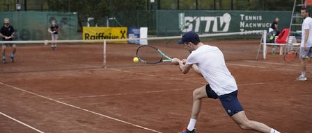 Ein Tennisspieler schwingt bei einem Match auf einem Sandplatz nach einem Ball. Er trägt ein weißes T-Shirt, blaue Shorts und Turnschuhe. Der Platz hat weiße Linien.