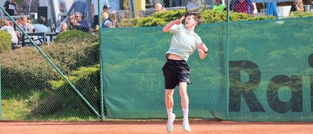 Ein Tennisspieler springt in der Luft und hält einen Schläger in der Hand, auf einem Sandplatz. Dahinter ist eine grüne Mauer mit 'RO' sichtbar. Zuschauer sitzen im Hintergrund und beobachten das Spiel.