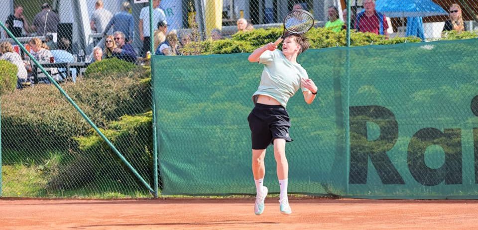 Ein Tennisspieler springt in der Luft und hält einen Schläger in der Hand, auf einem Sandplatz. Dahinter ist eine grüne Mauer mit 'RO' sichtbar. Zuschauer sitzen im Hintergrund und beobachten das Spiel.