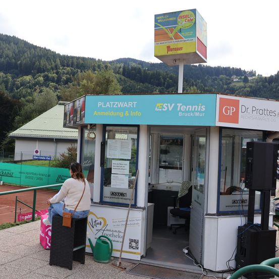 Eine Frau sitzt auf einer Bank vor einer Tickethütte auf einem Tennisplatz. Hinter der Hütte befindet sich ein Schild mit der Aufschrift Platzwart Anmeldung & Info und SVV Tennis. Im Hintergrund ist ein Tennisplatz zu sehen.