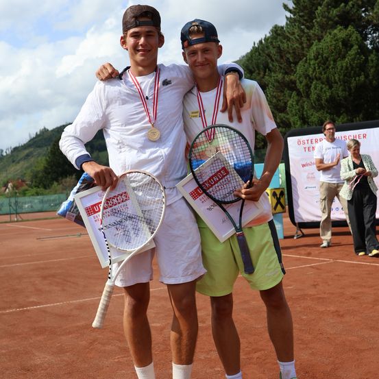 Zwei Männer stehen auf einem Tennisplatz, beide tragen Medaillen und halten Tennisschläger. Sie lächeln und posieren für ein Foto. Hinter ihnen steht ein Banner mit Text und Logos. Im Hintergrund stehen zwei Personen in der Nähe des Banners.