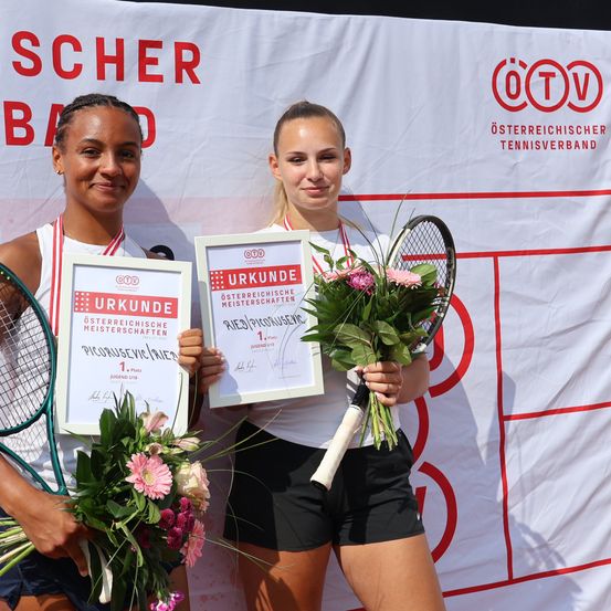 Zwei Tennisspielerinnen stehen nebeneinander, lächeln und halten Trophäen und Blumen. Der Hintergrund ist ein Banner mit rotem Text und Logos.