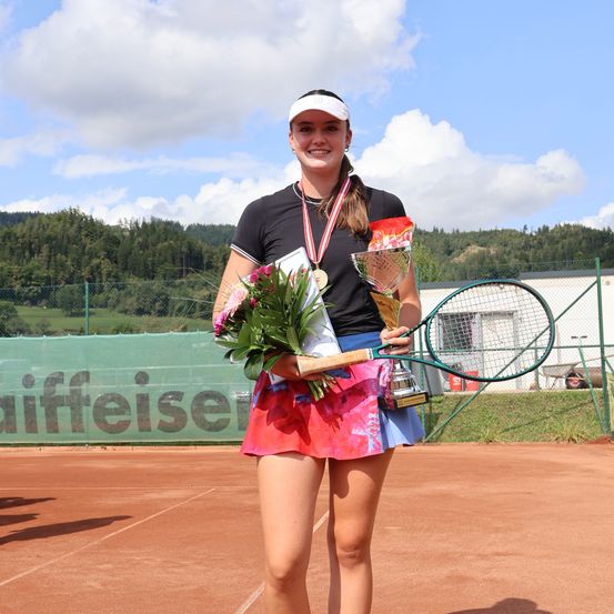 Eine junge Frau steht auf einem Tennisplatz und lächelt. Sie hält einen Blumenstrauß, einen Tennisschläger und einen Pokal. Hinter ihr steht ein Zaun mit dem Wort 'Waffeleiser' und einem Gebäude. Im Hintergrund sind Bäume und Berge zu sehen.
