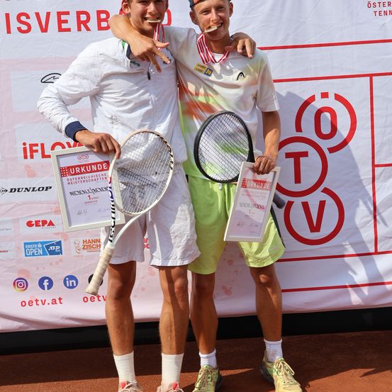 Zwei Männer stehen zusammen und halten Tennisschläger und Urkunden. Sie haben ihre Arme umeinander gelegt und lächeln. Sie tragen Tennisoutfits.