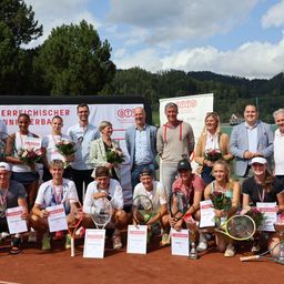 Eine Gruppe von Menschen posiert auf einem Tennisplatz für ein Foto. Sie halten Tennisschläger und einige tragen Medaillen. Hinter ihnen befindet sich ein Banner mit Text.