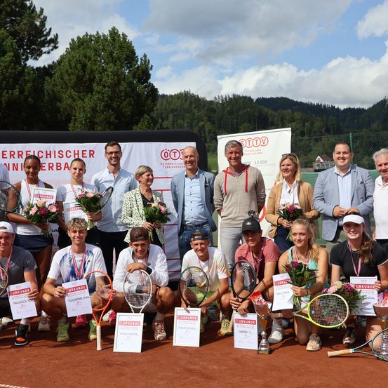 Eine Gruppe von Menschen posiert auf einem Tennisplatz für ein Foto. Sie halten Tennisschläger und einige tragen Medaillen. Hinter ihnen befindet sich ein Banner mit Text.