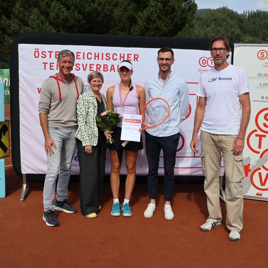 Fünf Personen posieren auf einem Tennisplatz mit Hintergrund. Eine Frau hält einen Blumenstrauß und einen Preis. Das Banner trägt die Aufschrift "Osterreichischer Tennisverband."