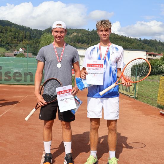 Zwei junge Männer stehen auf einem Tennisplatz, halten Tennisschläger und Urkunden in den Händen, mit Bergen und einem Zaun im Hintergrund.