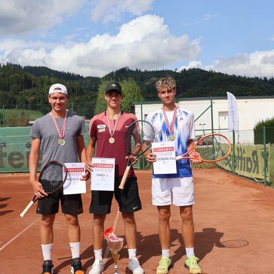 Drei Männer stehen auf einem Tennisplatz, halten Trophäen und Tennisschläger. Sie haben Medaillen um den Hals. Sie scheinen einen Sieg zu feiern. Hinter ihnen steht ein Gebäude und ein Zaun.