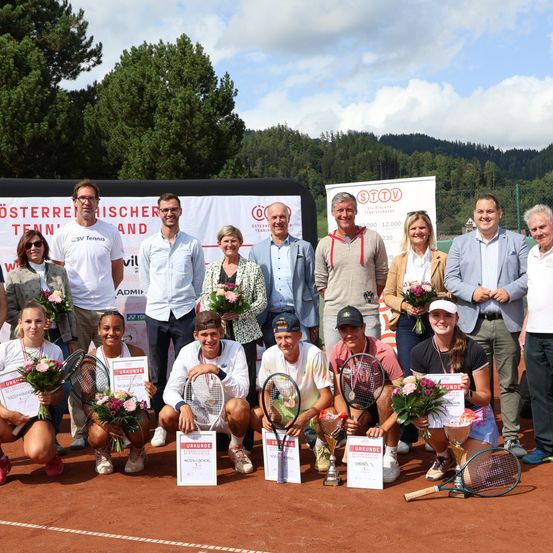 Eine Gruppe von Tennisspielern posiert für ein Foto auf einem Tennisplatz mit einem Banner im Hintergrund, trägt Auszeichnungen und hält Blumen und Trophäen.