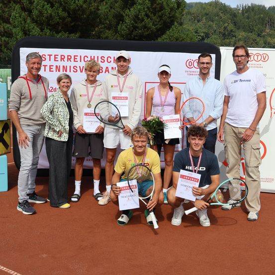 Eine Gruppe von Tennisspielern und Offiziellen posiert auf einem Tennisplatz mit Bannern im Hintergrund und hält Pokale und Tennisschläger in den Händen.