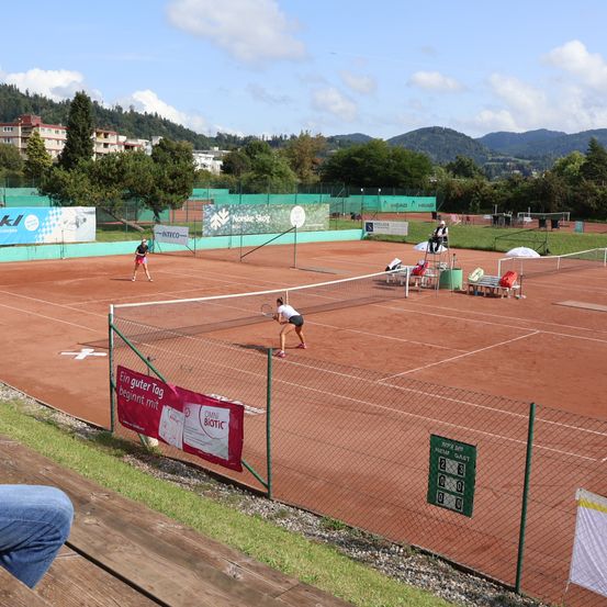 Ein Tennisplatz mit Spielern, Bannern und Zuschauern. Der Spielstand ist 0-0, und das Wetter ist sonnig.
