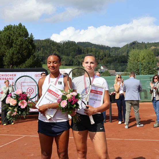 Zwei Frauen, die Tennisschläger und Blumensträuße halten, stehen auf einem Tennisplatz und lächeln für ein Foto. Hinter ihnen klatschen Menschen, und ein Zaun umgibt den Platz. Berge und Bäume sind im Hintergrund.