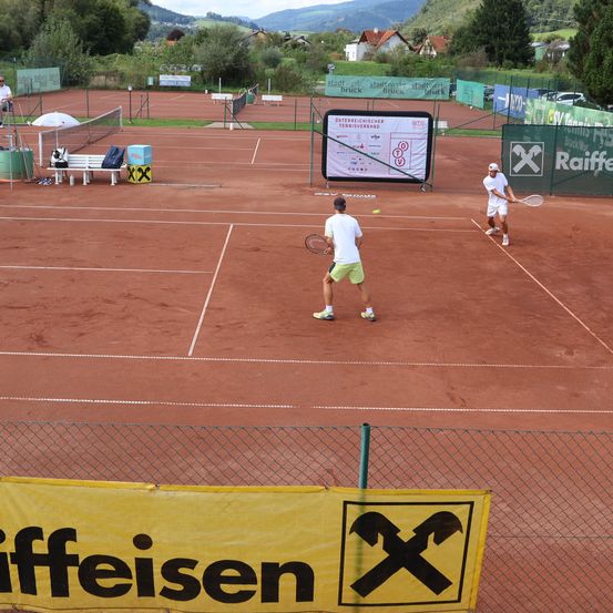 Zwei Männer spielen Tennis auf einem Sandplatz. Ein Spielstand und ein Sponsorenbanner sind sichtbar. Ein Mann beobachtet vom Stuhl aus.