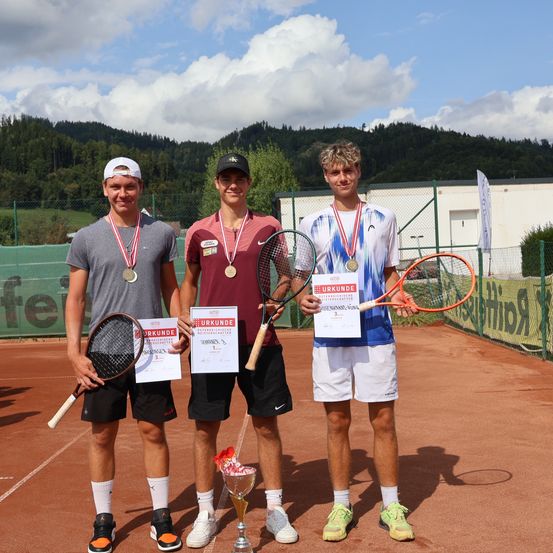 Drei Männer in Tenniskleidung stehen auf einem Platz mit Medaillen um den Hals. Sie halten Tennisschläger und Auszeichnungen. Ein Gebäude mit einem Banner ist im Hintergrund.