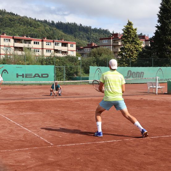Ein Tennisspieler bereitet sich darauf vor, einen Ball auf einem Sandplatz zu schlagen, während eine Person hinter ihm hockt. Der Platz ist von einem Zaun umgeben, mit HEAD- und Denn-Bannern. Im Hintergrund befinden sich Gebäude.