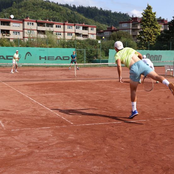Zwei Tennisspieler auf einem roten Platz, einer in Gelb und einer in Blau, spielen. Ein weiterer Spieler streckt sich im Vordergrund. Bäume und Gebäude im Hintergrund.