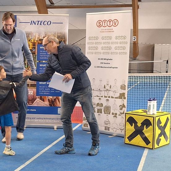 Drei Männer und ein Junge stehen auf einem blauen Platz mit einem Tennisnetz, Bannern und einem Kasten. Der Mann in der Mitte schüttelt dem Jungen die Hand.