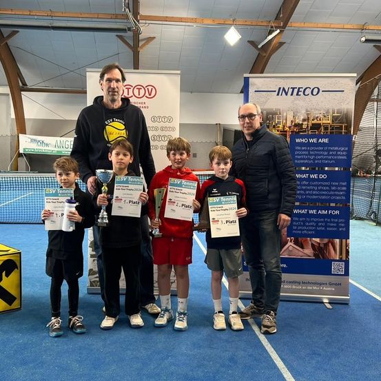 Fünf Jungen stehen in einer Reihe, halten Trophäen und Urkunden, lächeln. Dahinter stehen zwei Männer mit Bannern und einem Schild. Im Hintergrund ist ein Tennisplatz zu sehen.