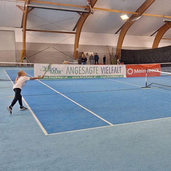 Ein junges Kind spielt Tennis auf einem Hallenplatz. Der Platz hat einen blauen Boden und weiße Markierungen. Ein Banner mit Text ist an der Wand hinter dem Spieler befestigt.