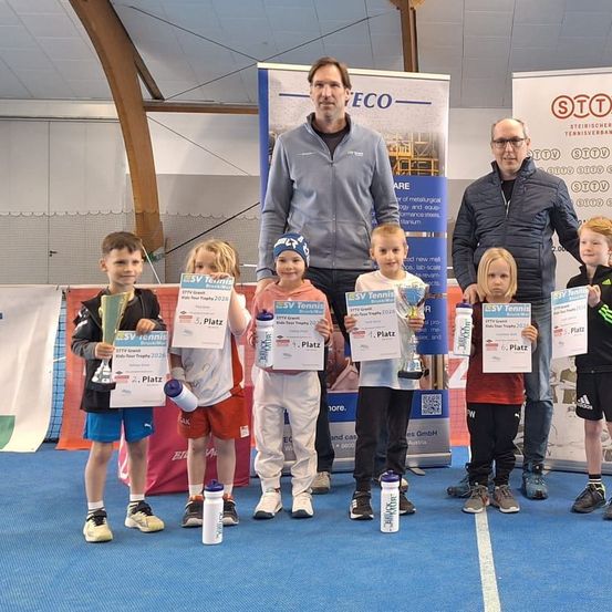 Eine Gruppe von Kindern steht mit Erwachsenen und hält Pokale und Urkunden. Sie sind in Sportkleidung gekleidet und stehen auf einem blauen Boden, wahrscheinlich nach einer Sportveranstaltung.