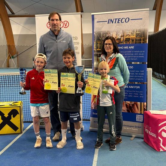 Drei junge Jungen und eine erwachsene Frau stehen hinter einem Tennisnetz und halten Pokale und Urkunden in den Händen. Die erwachsene Frau trägt eine Brille und die Jungen tragen Trikots und Shorts. Sie posieren wahrscheinlich für ein Foto nach einem Wettbewerb.