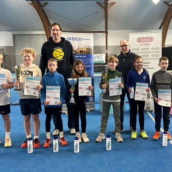 Eine Gruppe junger Tennisspieler steht auf einem Tennisplatz mit Pokalen und Urkunden in der Hand und zwei Männer hinter ihnen. Sie lächeln und posieren für ein Foto.