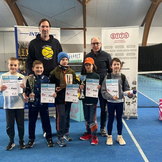 Eine Gruppe von Kindern steht auf einem Tennisplatz und hält Trophäen. Hinter ihnen stehen zwei Männer und Banner. Sie alle lächeln für ein Foto. Die Kinder tragen Jacken, Mützen und Turnschuhe. Der Boden ist blau.