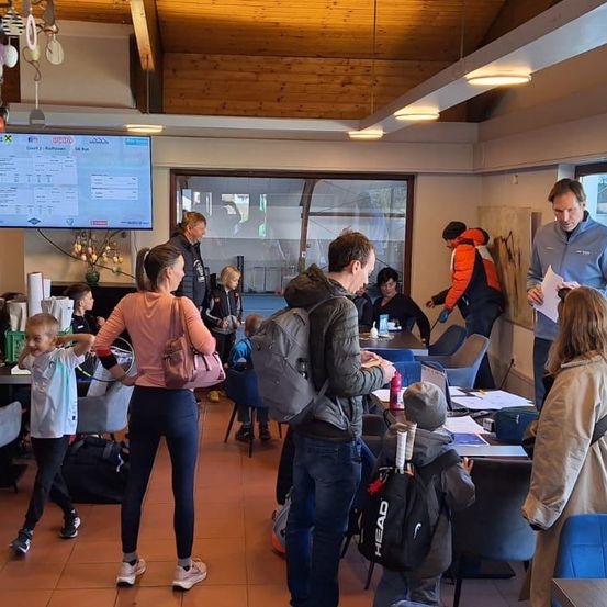 In einem Raum mit einer großen Leinwand versammeln sich Erwachsene und Kinder. Einige lesen Zeitungen, während andere sich unterhalten. Ein Mann hält einen Tennisschläger.