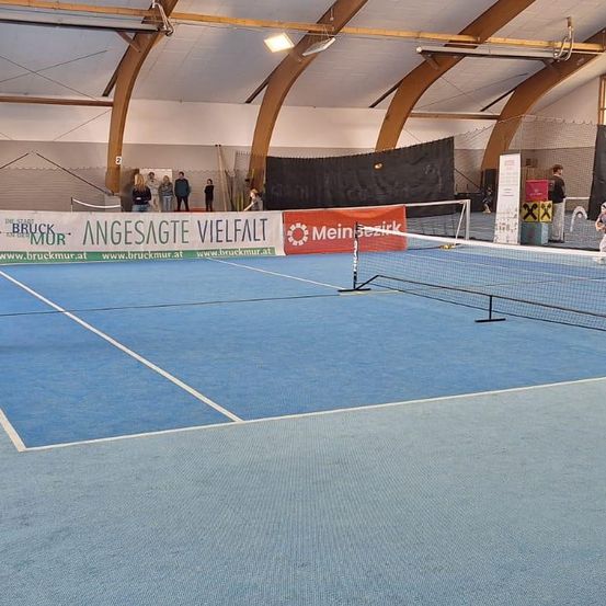 Ein Tennisplatz mit blauem Boden. Eine Banner mit Text an der Wand. Einige Leute stehen in der Nähe der Wand. Ein Netz ist in der Mitte des Platzes.