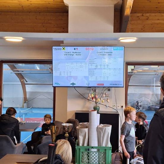 Sporthalle mit Holzdecken und Glasfenstern, ein großer Bildschirm zeigt Ergebnisse an, Trophäen auf einem Tisch, und Menschen beobachten.