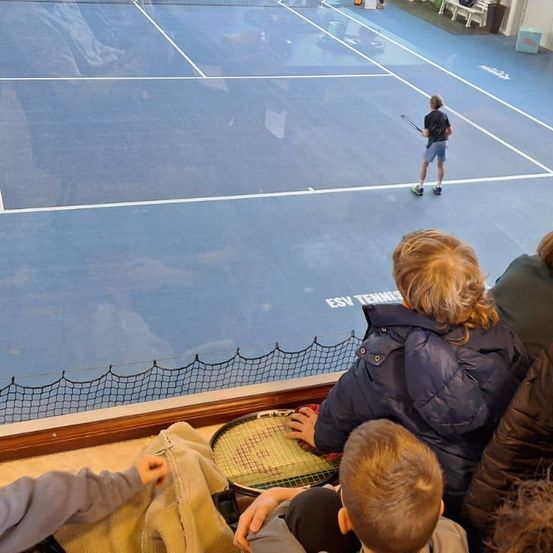 Ein Tennisplatz mit einem Jungen, der einen Schläger hält, ist zu sehen. Zuschauer beobachten von den Tribünen aus. Ein Netz und das Wort 'ESV' sind auf dem Platz sichtbar.
