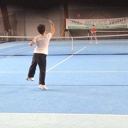 Zwei Jungen spielen Tennis auf einem blauen Platz. Ein Junge serviert, während der andere sich auf den Rückschlag vorbereitet. Ein Banner, das für 'Bruckmur' wirbt, ist im Hintergrund sichtbar.