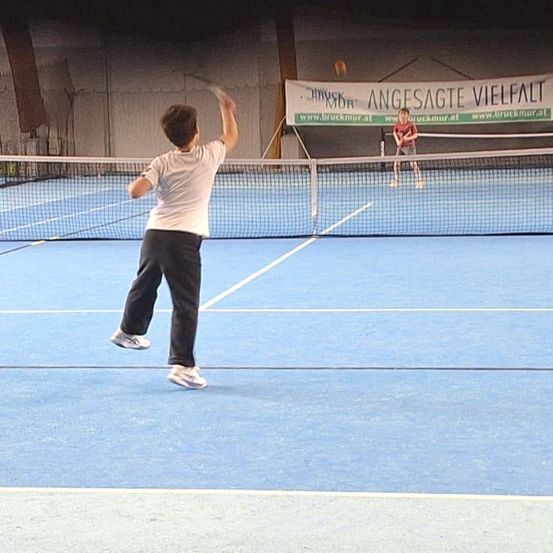 Zwei Jungen spielen Tennis auf einem blauen Platz. Ein Junge serviert, während der andere sich auf den Rückschlag vorbereitet. Ein Banner, das für 'Bruckmur' wirbt, ist im Hintergrund sichtbar.