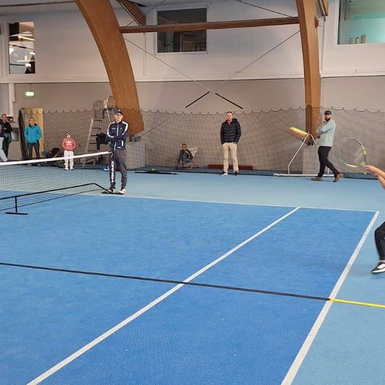 Mehrere Leute spielen Tennis in einer Halle. Ein Mann serviert. Andere stehen oder sitzen. Eine Netzmitte befindet sich in der Mitte.
