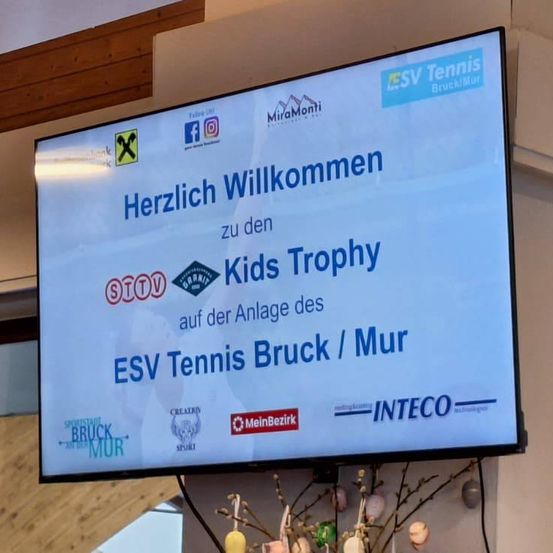 Ein Bildschirm zeigt eine Willkommensbotschaft für den Kids Trophy bei ESV Tennis Bruck/Mur. Verschiedene Sponsorenlogos sind sichtbar.