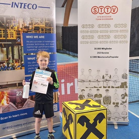 Ein junger Junge steht mit einem Zertifikat und einem Becher in der Hand und lächelt vor einem Tennisbanner und einem Kasten. Hinter ihm ist ein Banner, das ein Unternehmen bewirbt, mit Bildern von Arbeitern und Ausrüstung.