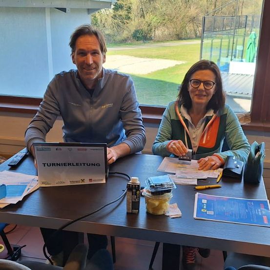 Zwei Personen sitzen an einem Tisch mit einem Laptop und anderen Gegenständen. Ein Mann lächelt und eine Frau schaut auf eine Karte. Papiere, ein Stift und eine Fernbedienung sind auf dem Tisch. Ein Fenster mit Blick auf ein grasbewachsenes Feld ist hinter ihnen.