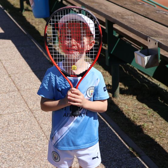 Ein junger Junge mit einem Manchester City Trikot hält einen Tennisschläger neben einer Bank im Freien.