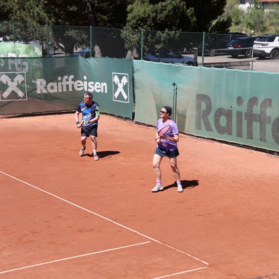 Zwei Personen spielen Tennis auf einem Platz mit einem Zaun, der mit Raiffeisen-Markenwerbung versehen ist. Ein Mann und eine Frau, beide halten Tennisschläger, befinden sich auf gegenüberliegenden Seiten.