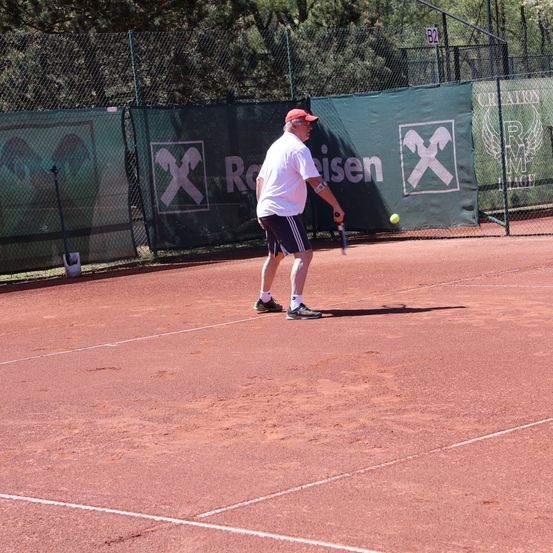 Ein älterer Mann in Hut und Turnschuhen spielt Tennis auf einem Sandplatz, mit einem Ball in der Luft. Hinter ihm ist eine grüne Umzäunung mit Logos und einem Schild sichtbar.