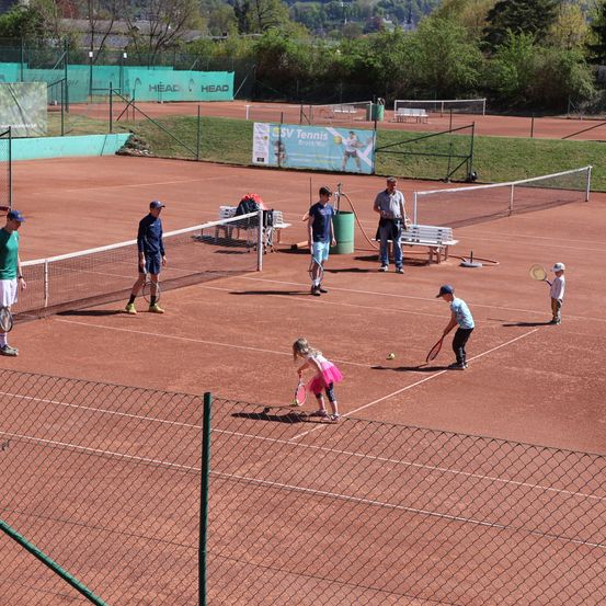 Menschen spielen Tennis auf einem Außenplatz. Mehrere Spieler, darunter Kinder, sind beim Spiel zu sehen. In der Nähe befinden sich Bänke und ein Mülleimer.